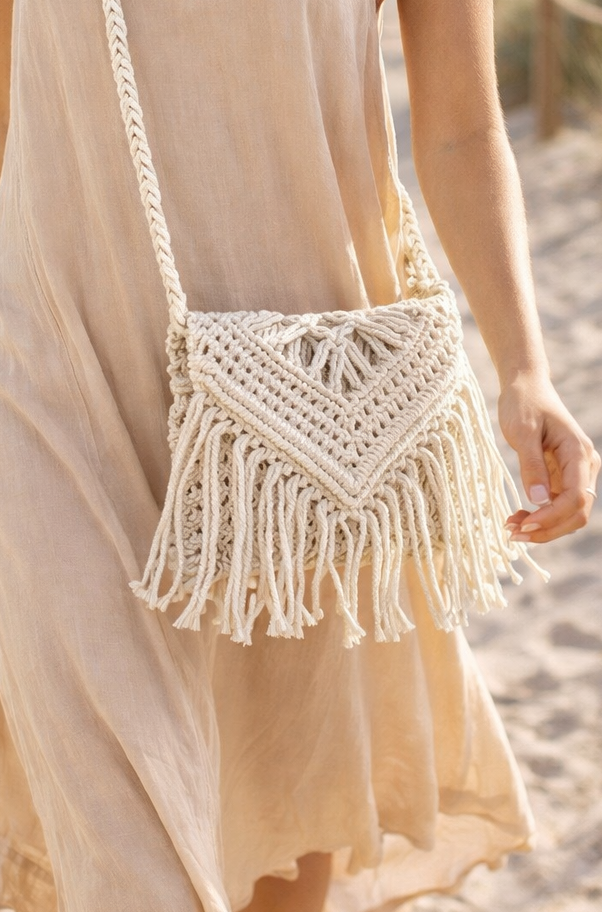Model wearing an ivory macramé crossbody bag with fringe on a sandy beach